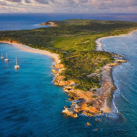 Aerial view of a lush, green island with sandy beaches, surrounded by turquoise water, and sailboats.