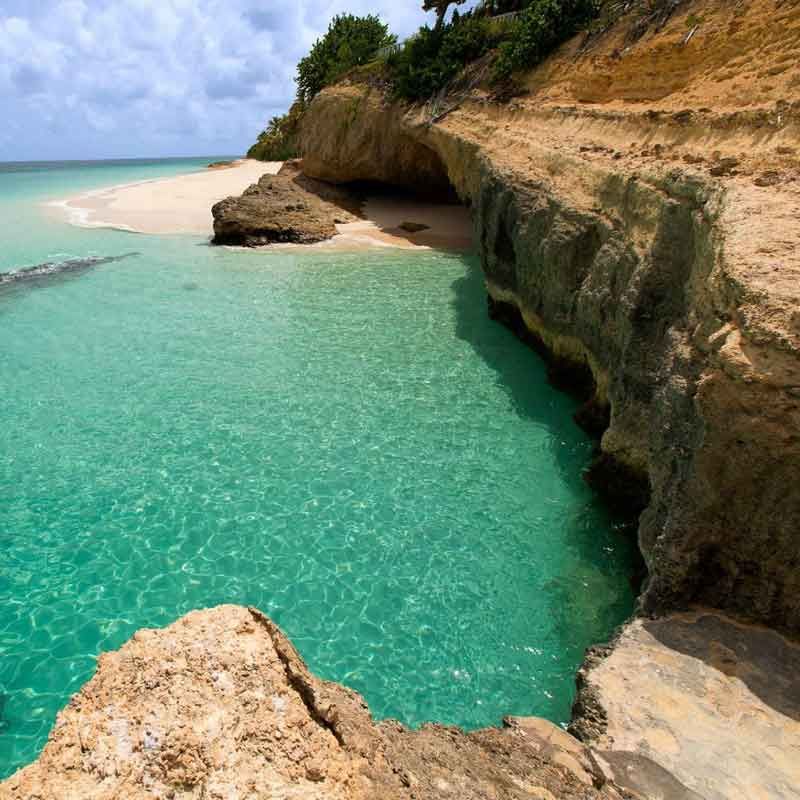 Turquoise water laps against a sandy beach, bordered by tan cliffs and a cave.