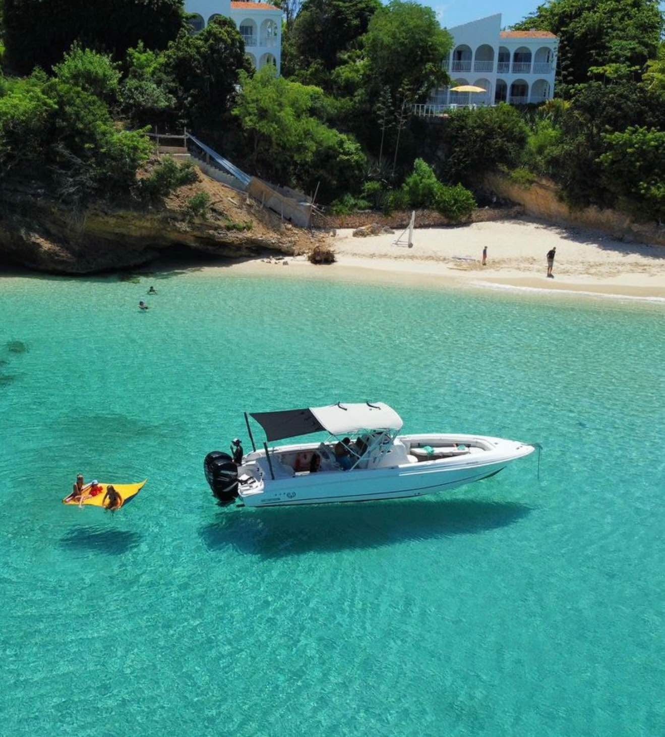 Boat in clear turquoise water near a white sand beach and lush green hillside with buildings.