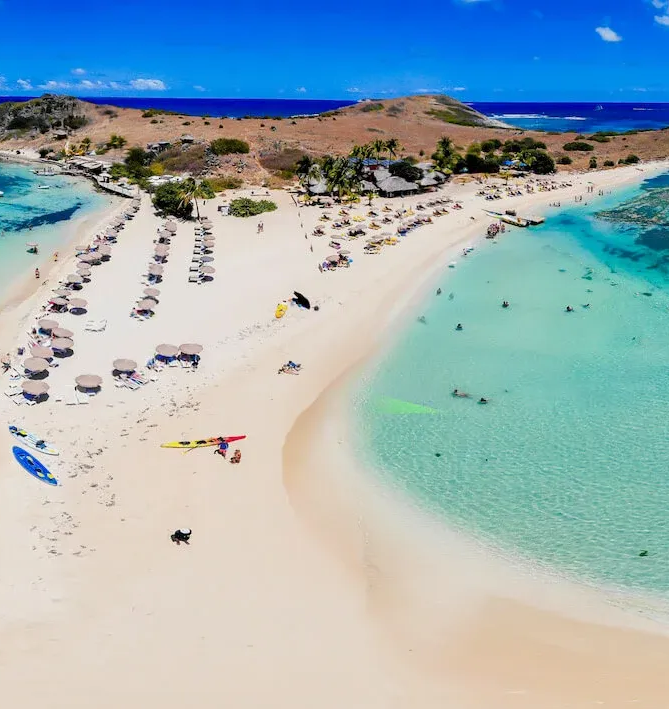 Aerial view of a white sand beach with clear turquoise water and people swimming and sunbathing.