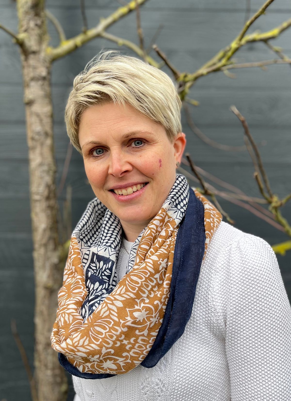 Eine Frau mit Schal und weißem Pullover lächelt vor einem Baum.