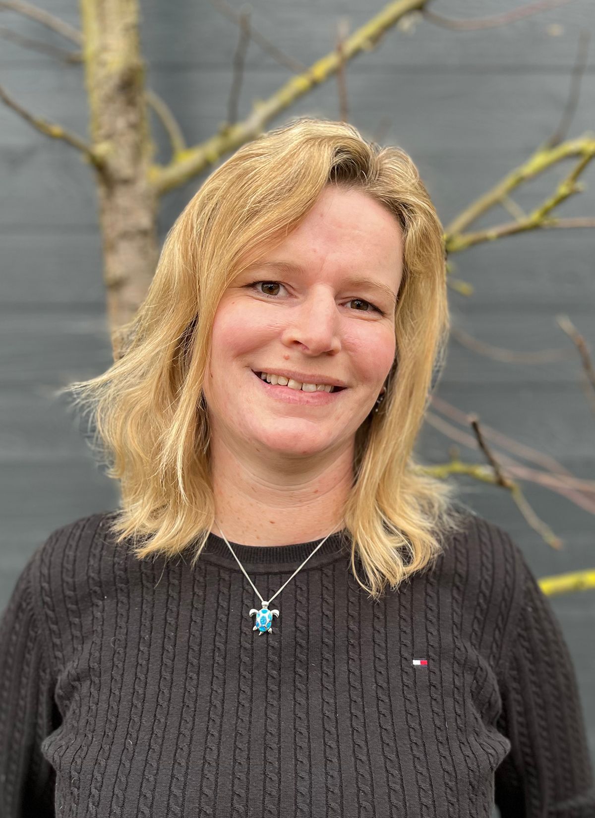Eine Frau mit einem schwarzen Pullover und einer Halskette lächelt vor einem Baum.