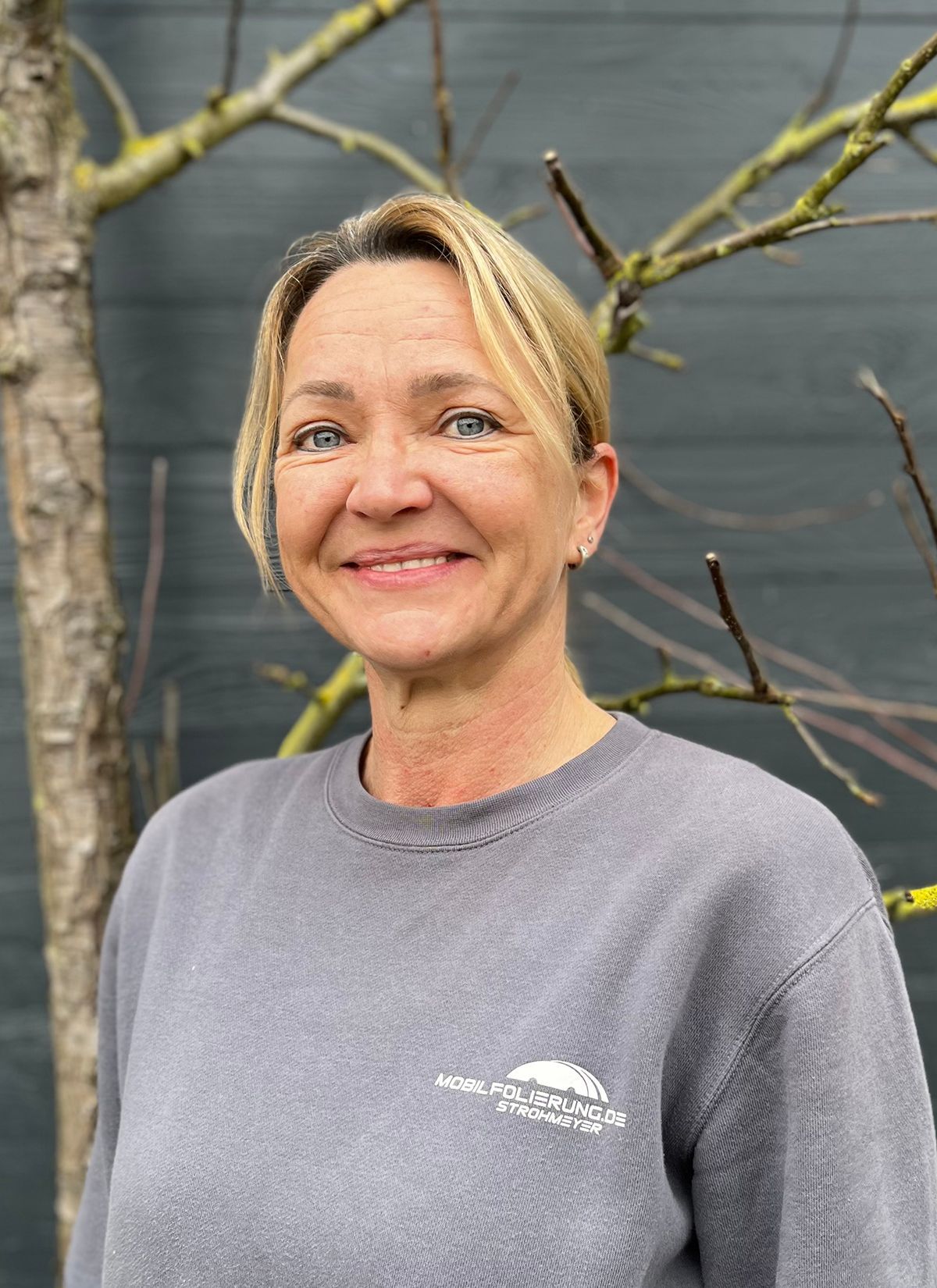 Eine Frau in einem grauen Pullover lächelt vor einem Baum.