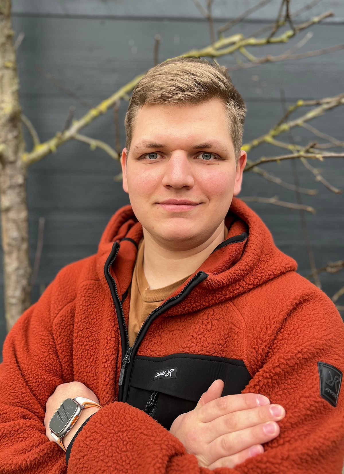 Ein junger Mann mit einer roten Jacke und einer Uhr steht mit verschränkten Armen da.