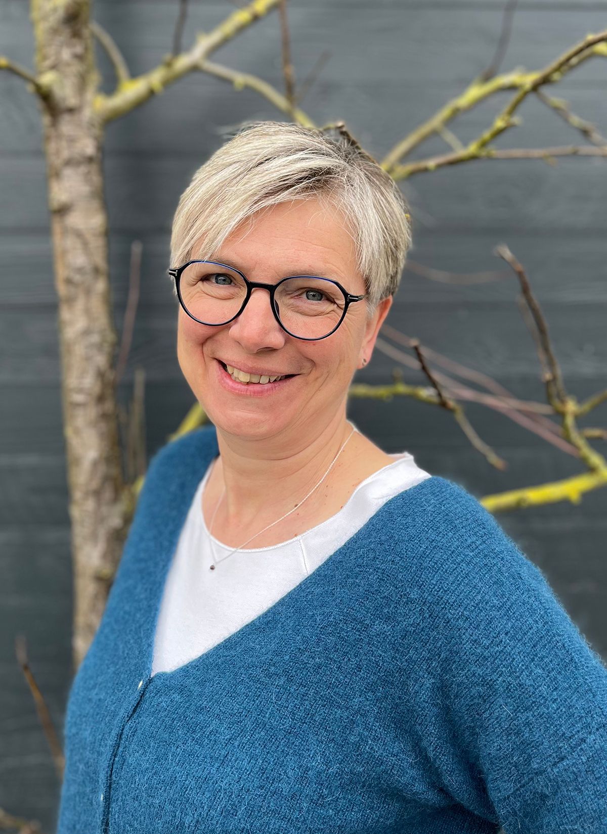 Eine Frau mit Brille und blauem Pullover lächelt vor einem Baum.