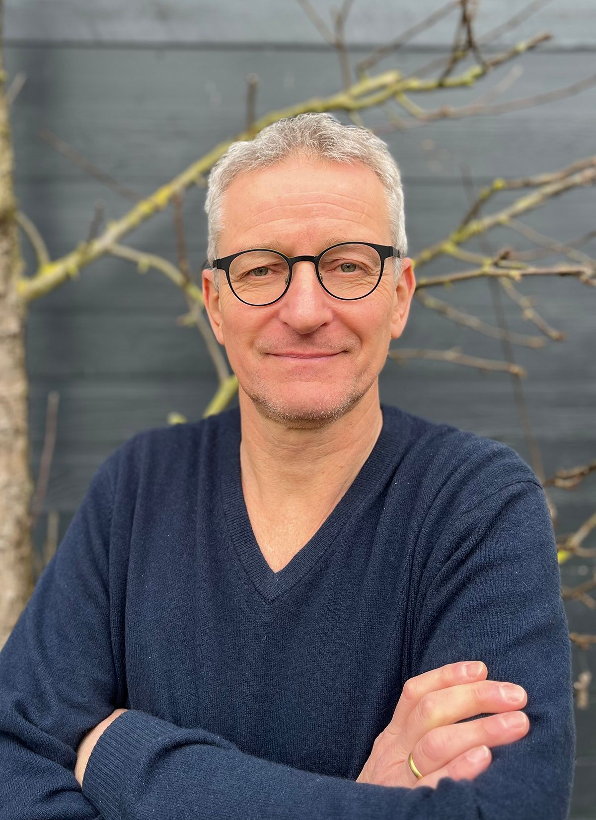 Ein Mann mit Brille und blauem Pullover steht mit verschränkten Armen da.