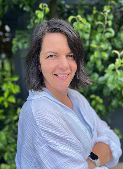 Frau mit dunklem Haar, lächelnd, trägt ein hellblaues Oberteil, die Arme verschränkt, vor grünem Laub.