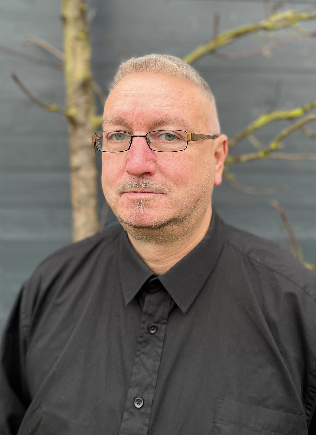 Ein Mann mit Brille und schwarzem Hemd steht vor einem Baum.