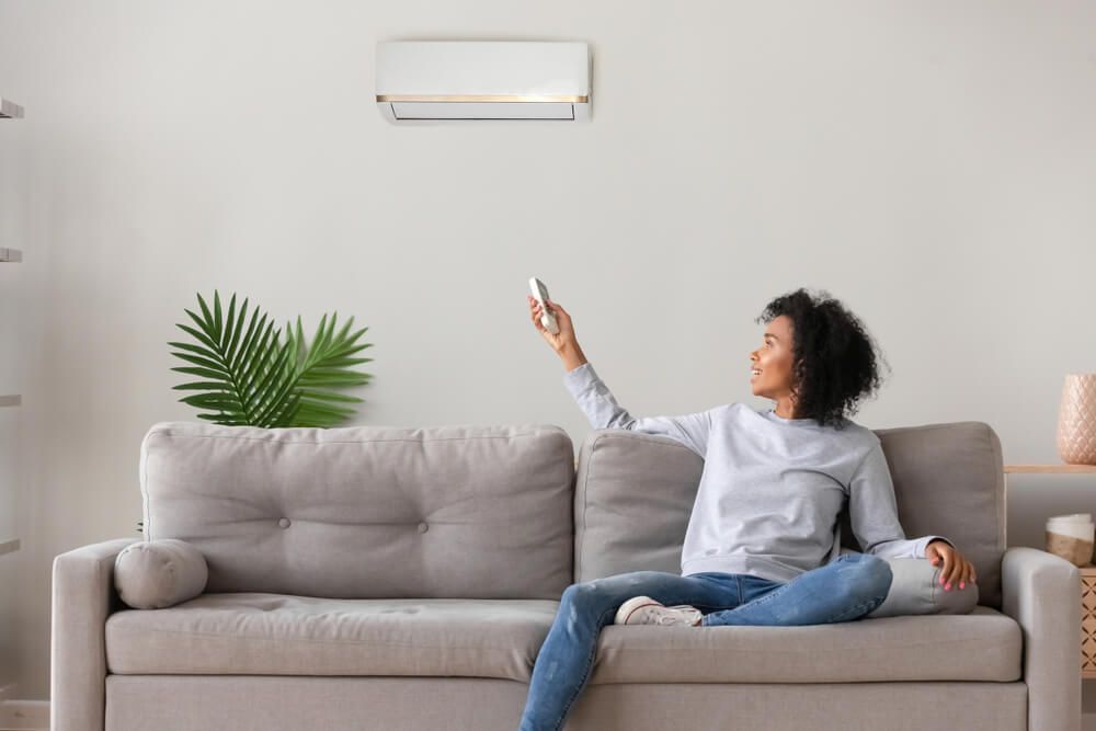 A Woman is Sitting on a Couch Holding a Remote Control of Air Condition — Refrigerated Technologies (QLD) Pty Ltd In Bowen, QLD