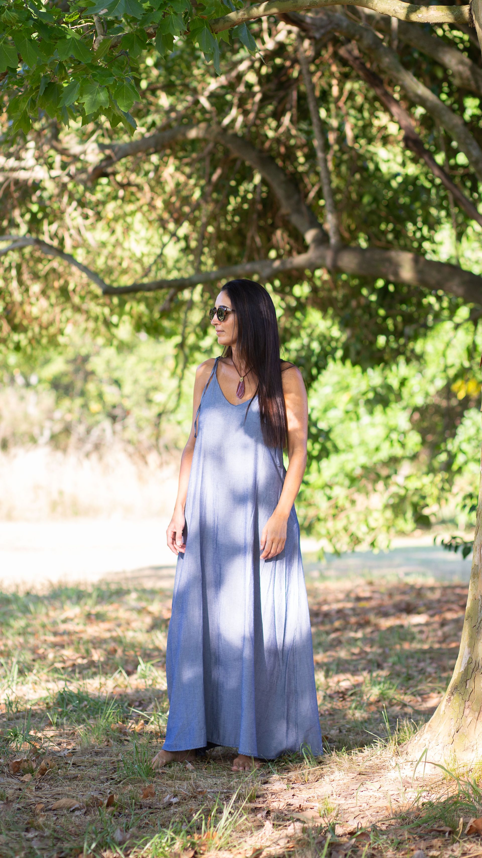 Mujer con vestido largo de rayas azules y blancas, gafas de sol, de pie al aire libre bajo los árboles.