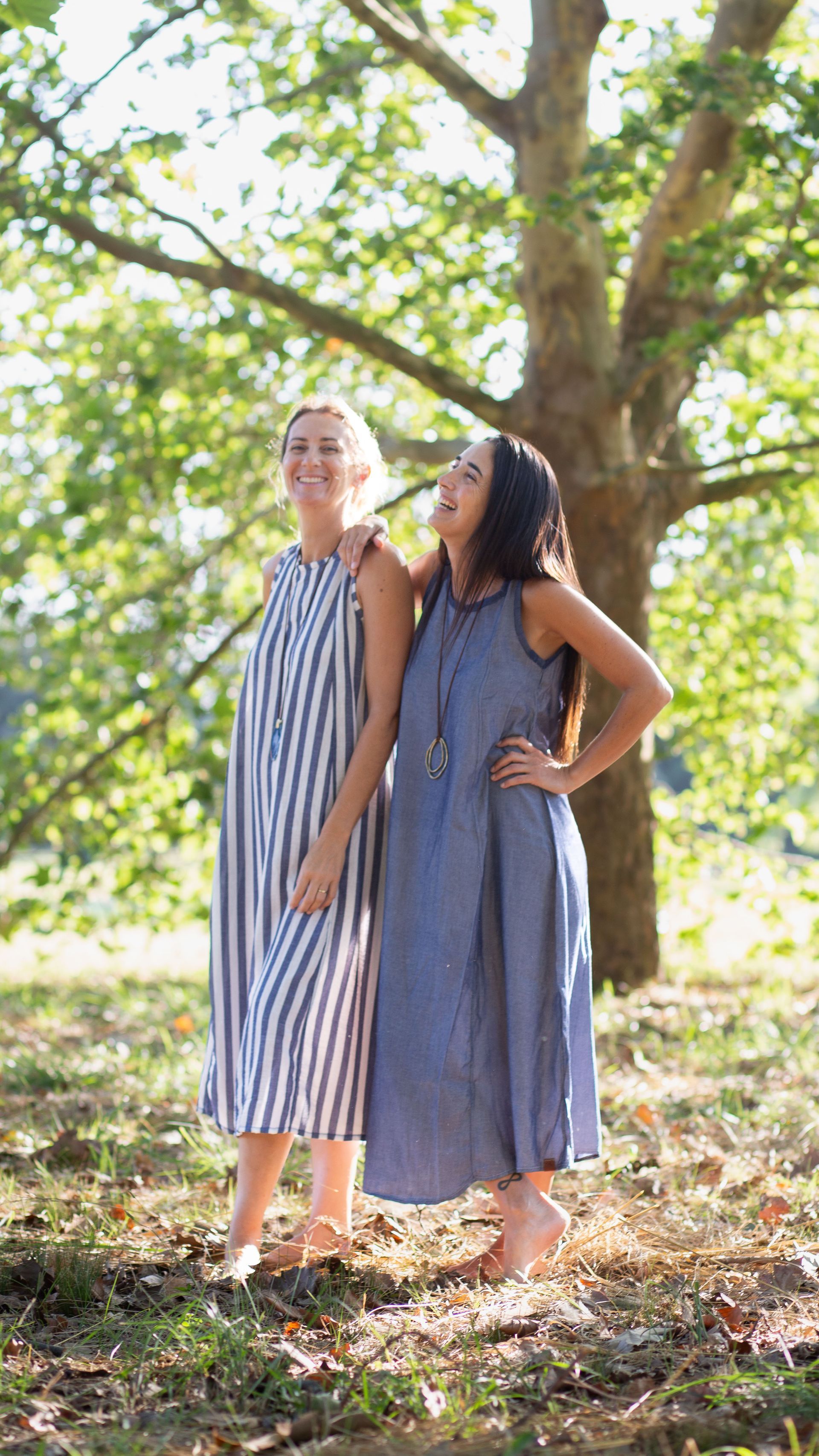 Dos mujeres, al aire libre, sonriendo; una con un vestido de rayas, otra con un vestido azul, de pie cerca de un árbol.