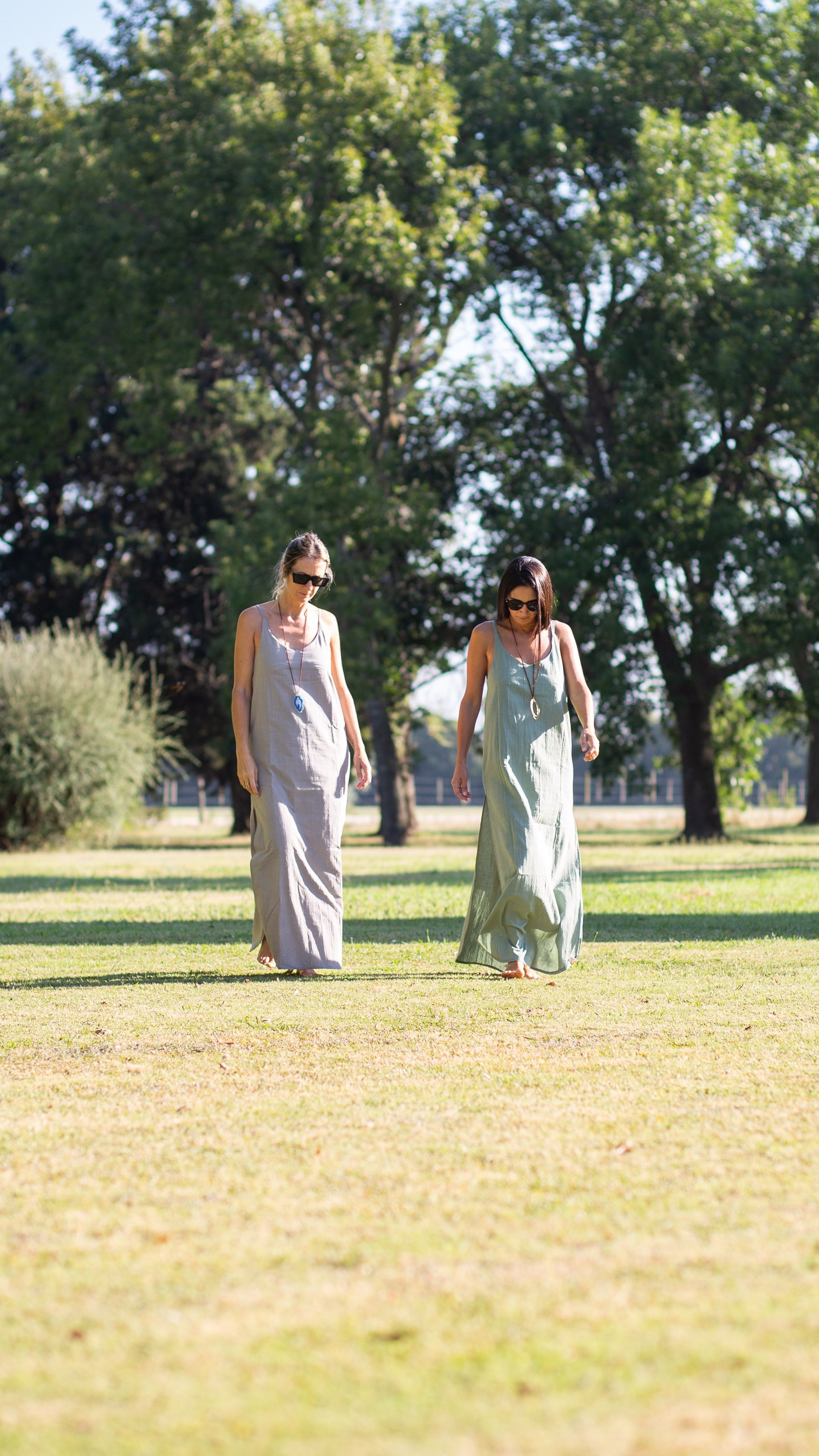 Dos personas con vestidos largos caminando por un campo de hierba con árboles al fondo.