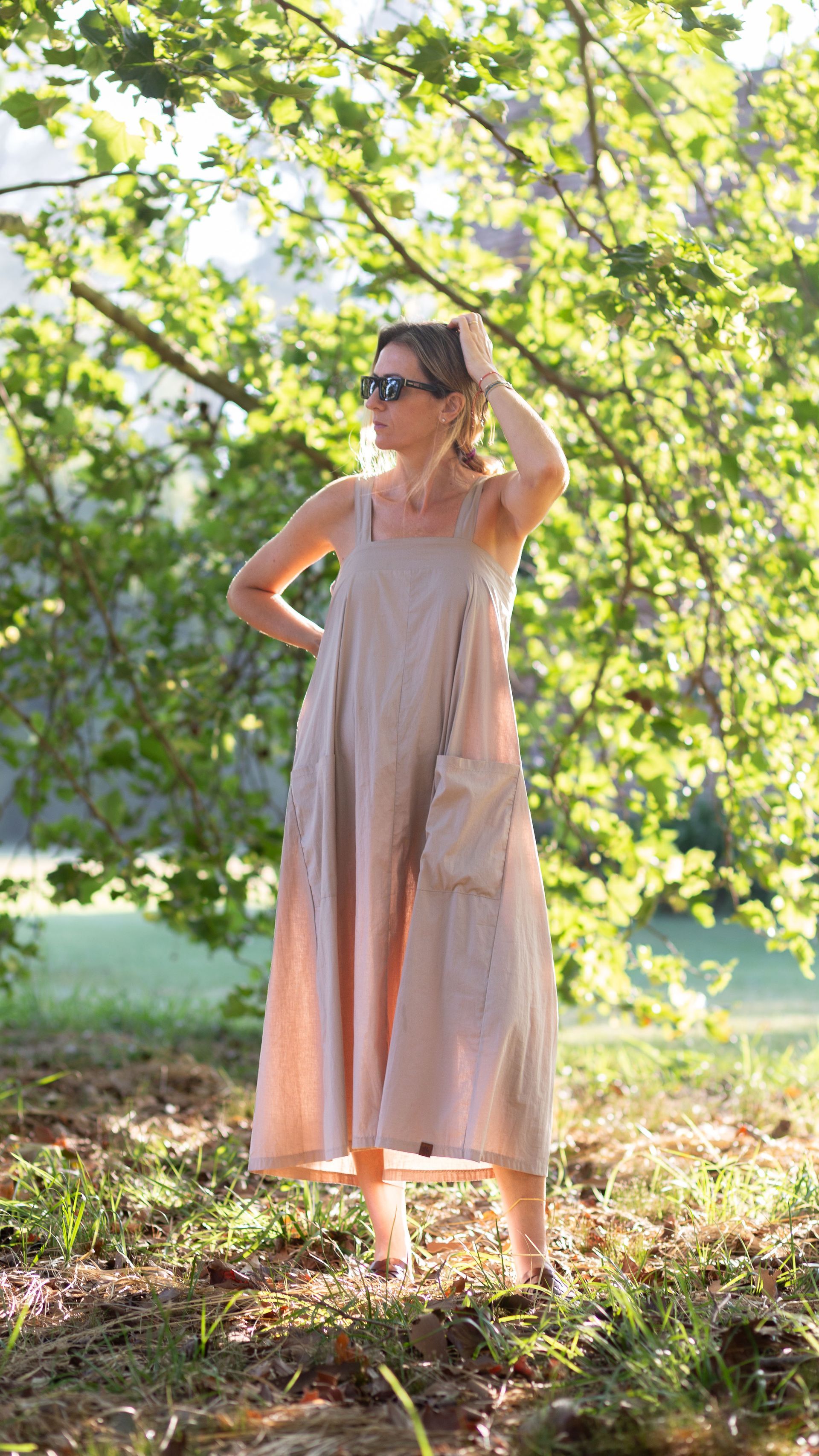 Mujer con un vestido rosa de pie descalza en un parque soleado, con la mano en el cabello.