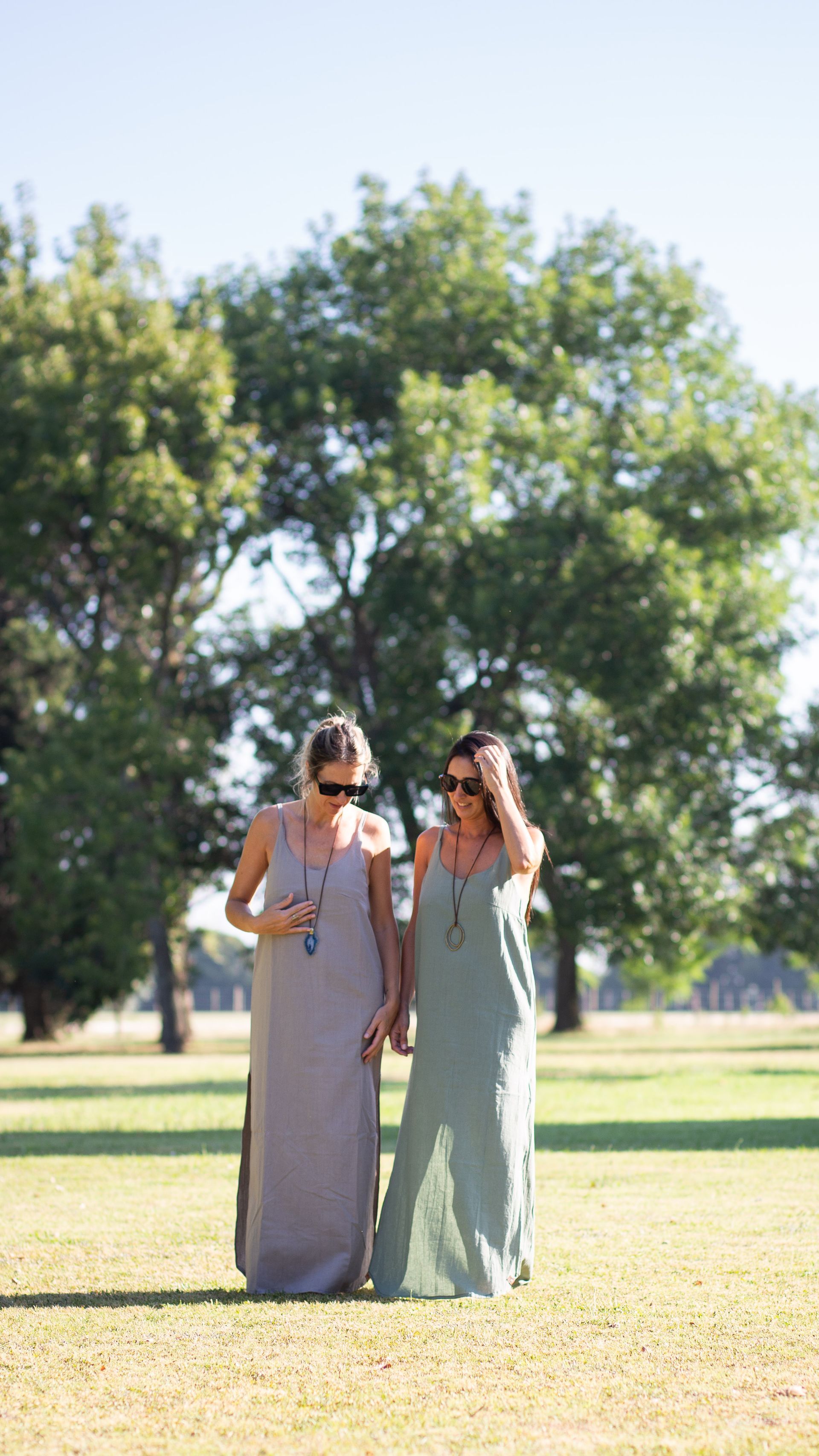 Dos personas con vestidos largos caminando por un campo de hierba con árboles al fondo.