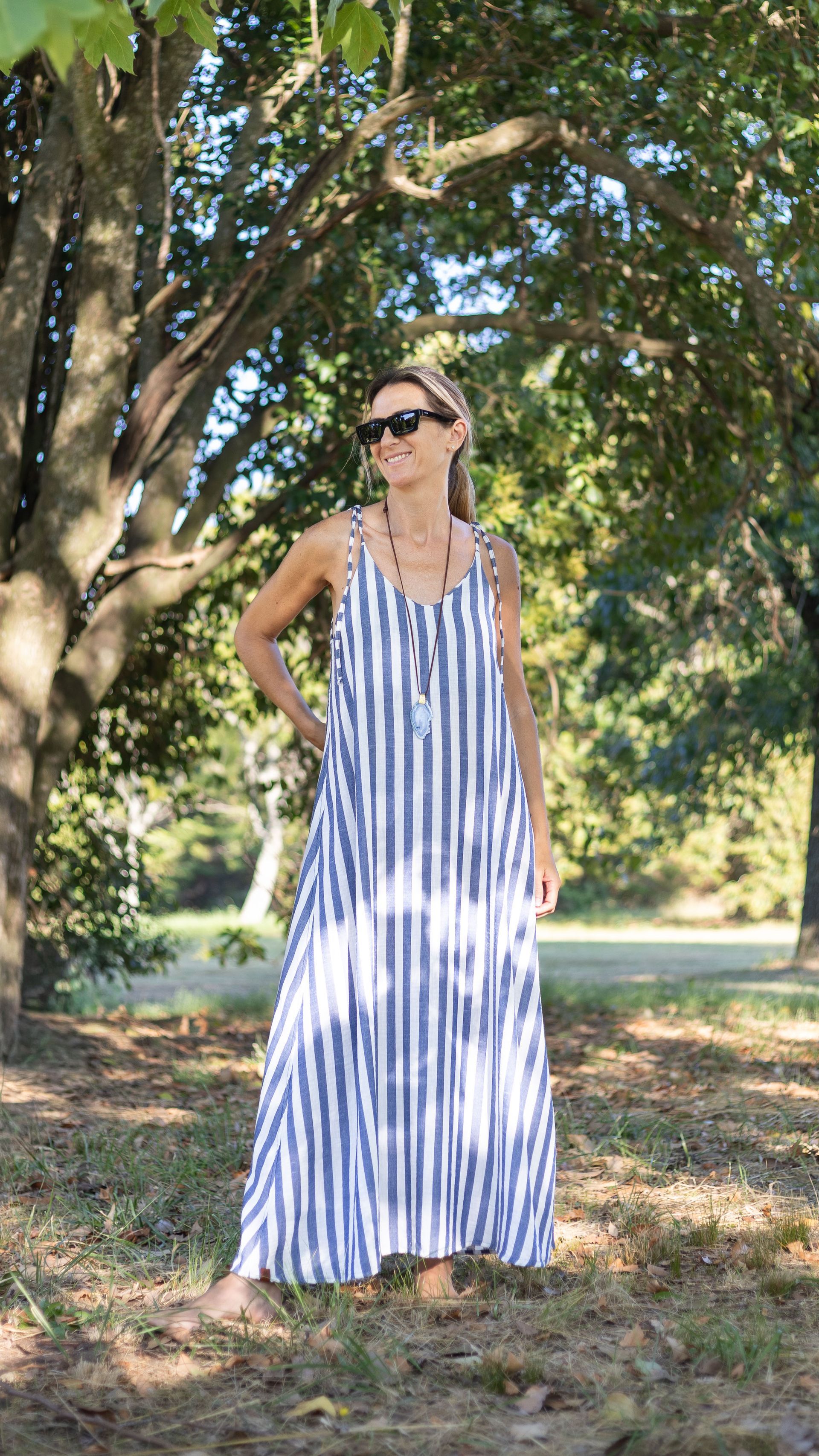 Mujer con vestido largo de rayas azules y blancas, gafas de sol, de pie al aire libre bajo los árboles.