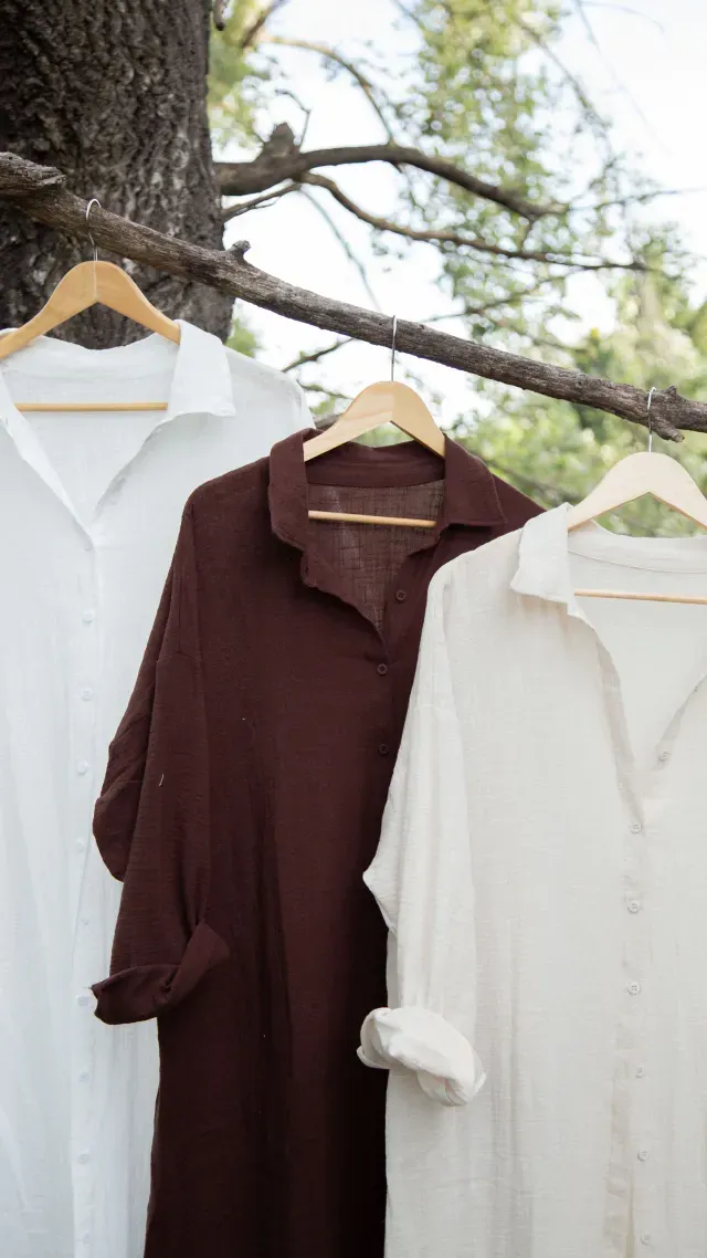Mujer vestida con una camisa blanca de manga larga, un vestido negro y sandalias de pie sobre el césped al aire libre.