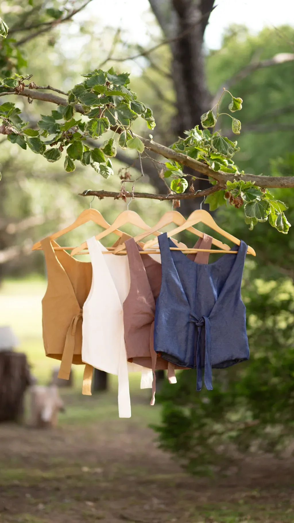 Cuatro chalecos de color marrón, crema, tostado y azul cuelgan de la rama de un árbol al aire libre.