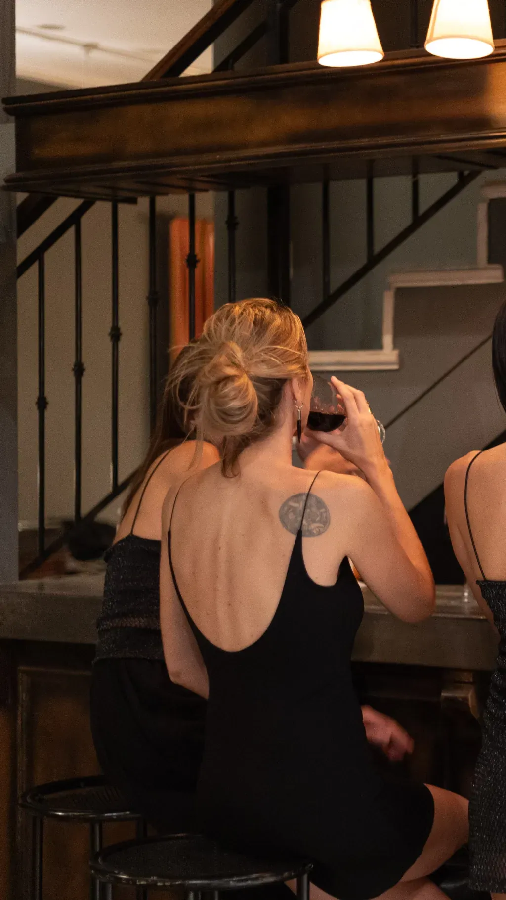 Una mujer vestida de negro y con un tatuaje en la espalda bebe de una copa de vino en un bar.