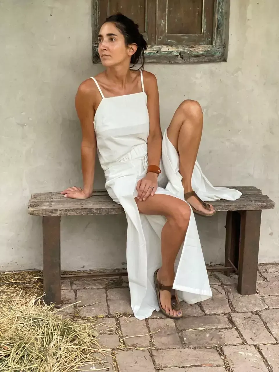 Una mujer vestida de blanco se sienta en un banco de madera desgastada, con las piernas cruzadas y mirando hacia un lado.