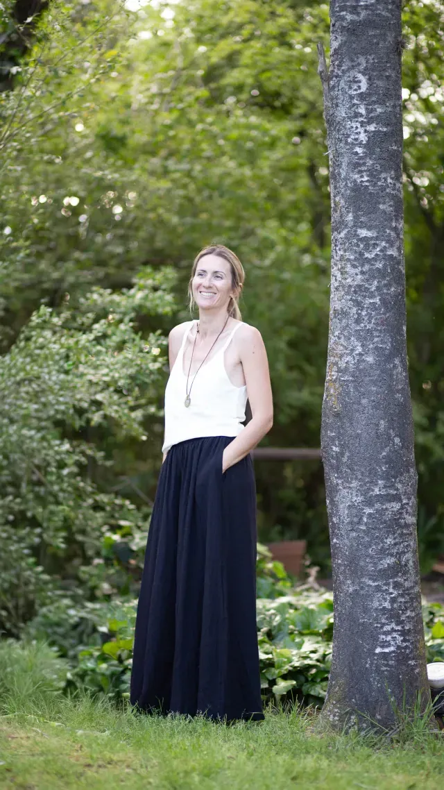 Mujer con top blanco y falda negra, parada afuera con la mano en el bolsillo, al lado de un árbol.