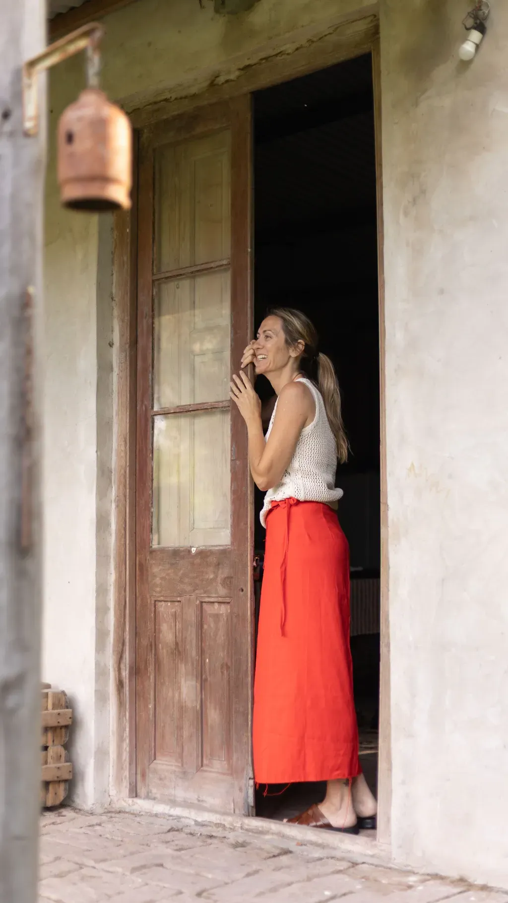 Mujer con falda roja y blusa blanca sonríe en un portal. Edificio rústico con decoración colgante.