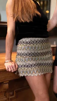 Mujer con top negro y falda estampada apoyada sobre mostrador de madera.