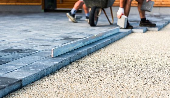 A man is pushing a wheelbarrow while another man lays bricks on a driveway.