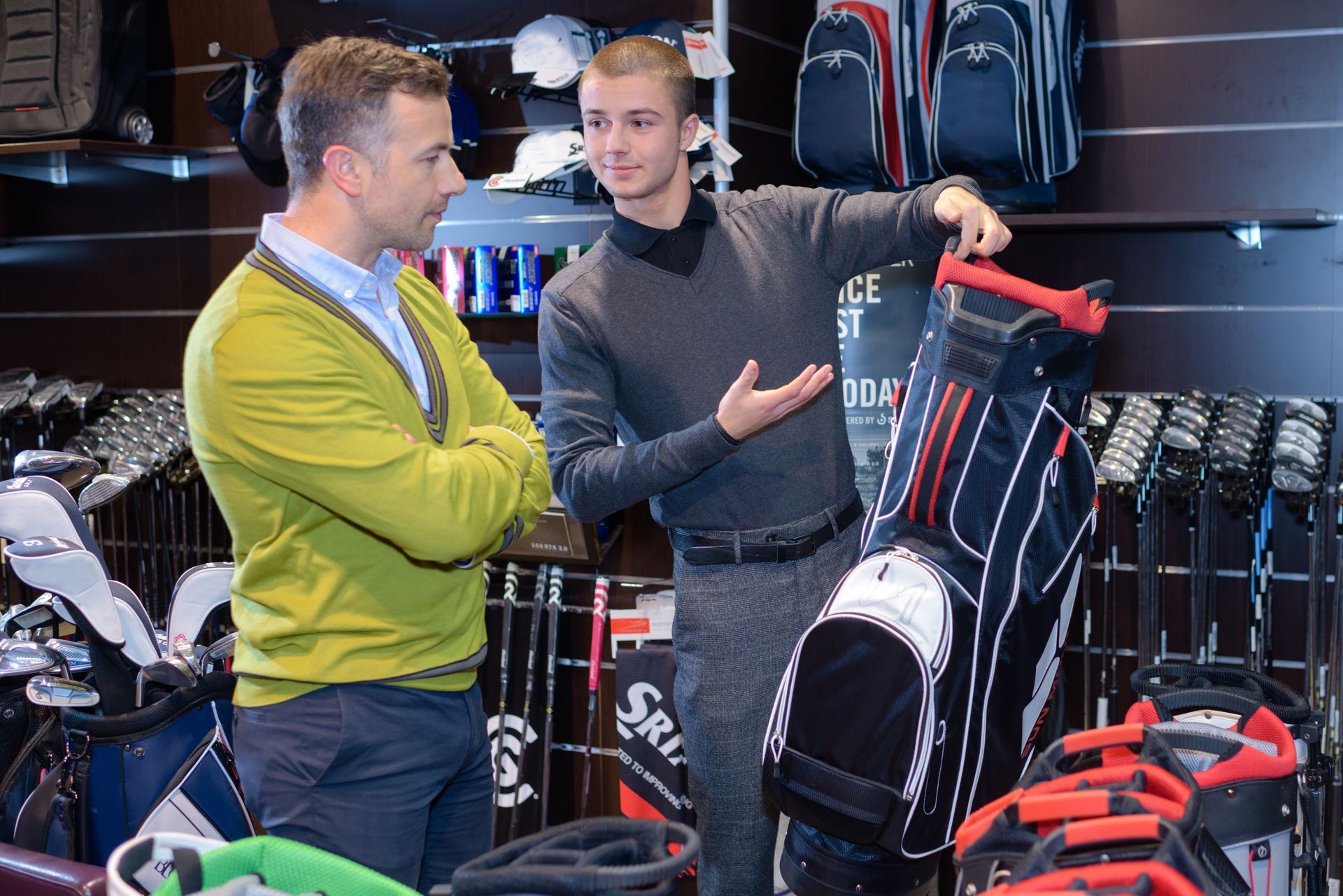 A golf store employee is assisting a customer with golf bag selection.