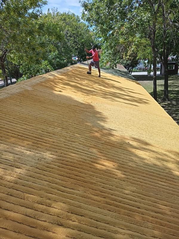 Persona aplicando aislamiento con espuma en aerosol sobre un techo amarillo, árboles en el fondo.