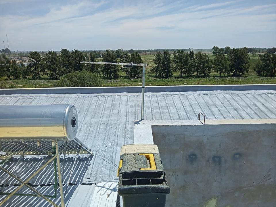 Escena en azotea con calentador de agua solar y antena. Campo verde con árboles al fondo.