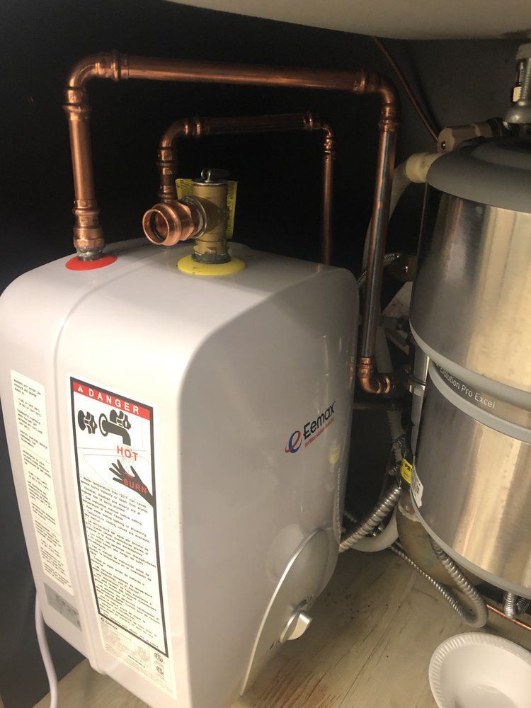 a white water heater is sitting under a sink with copper pipes .