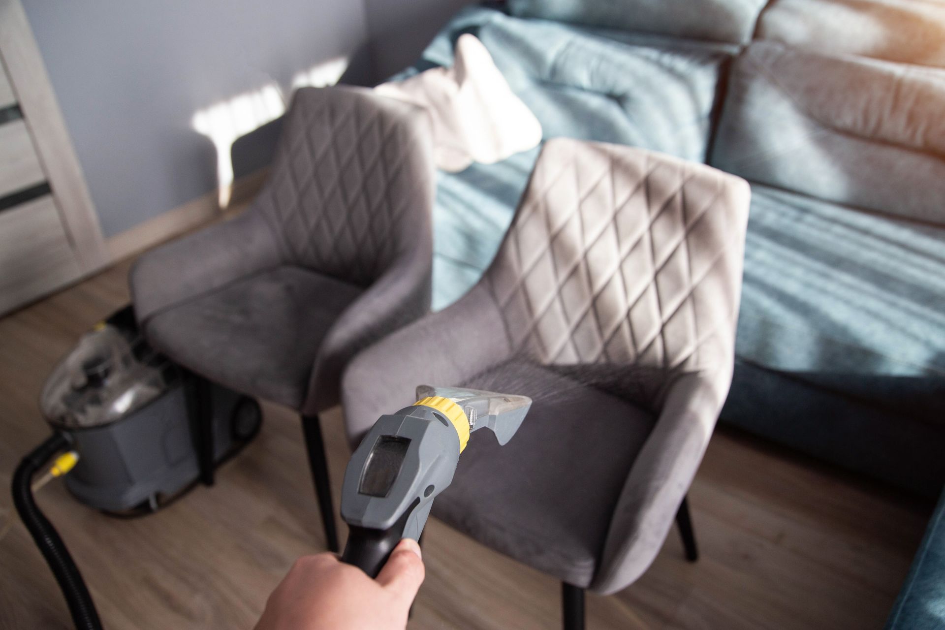 Person cleaning a gray upholstered chair with a handheld upholstery cleaner; sofa in background.