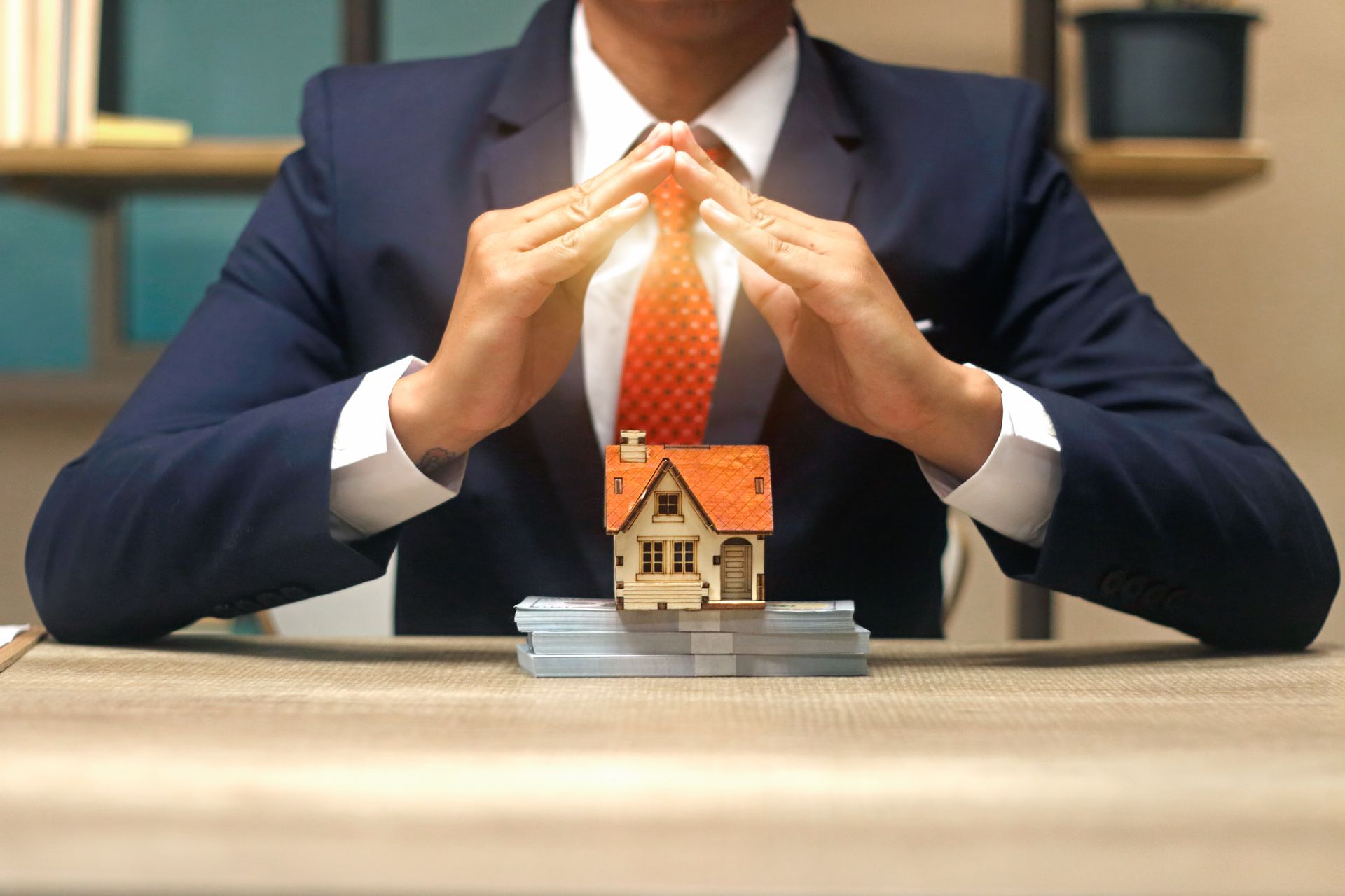 A man in a suit and tie is sitting at a table holding a model house. A man in a suit and tie is sitting at a table holding a model house.