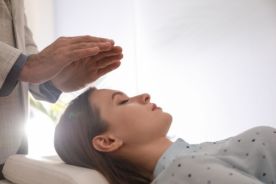 Hands over a woman's head during a reiki session; she lies with eyes closed on a massage table indoors.