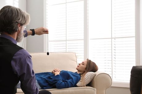 A person is hypnotizing another person on a couch with a swinging pendulum.