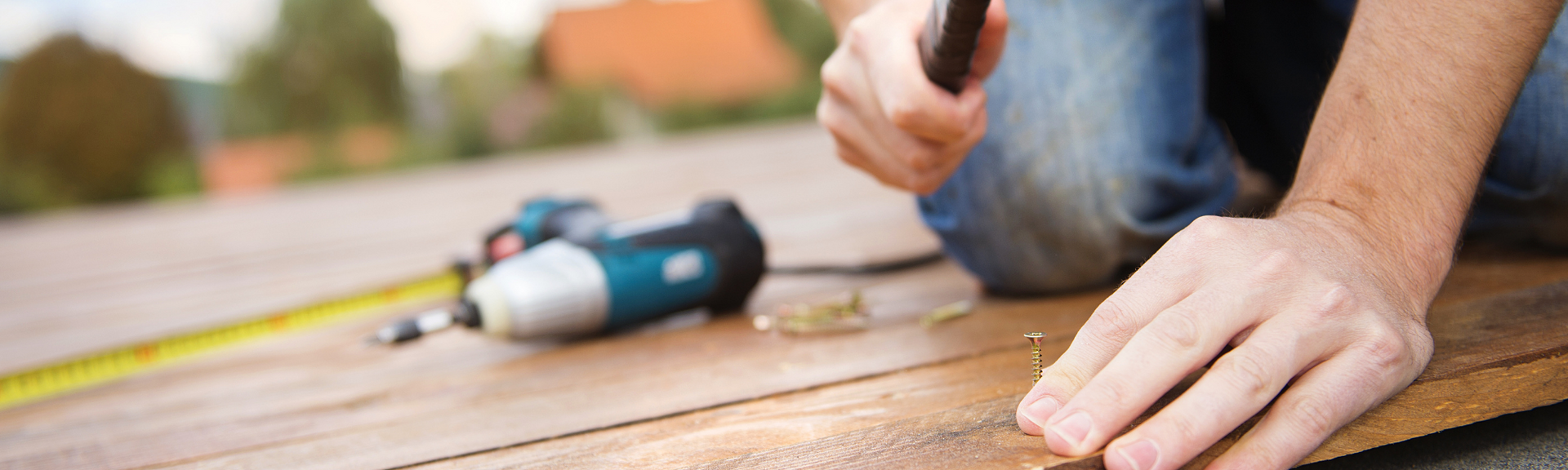 A person uses a hammer and a power drill to install wooden deck planks.