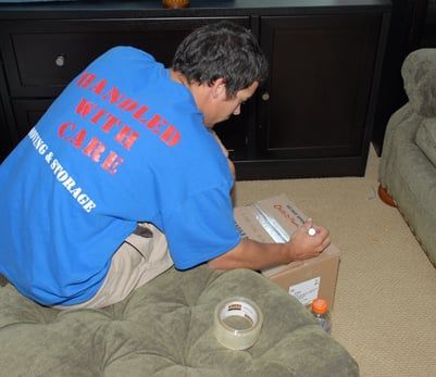 Man in blue shirt, kneeling, taping a cardboard box on a green cushion.