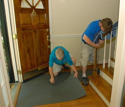 Two men installing a gray floor mat in a home entryway. One kneels, the other leans over a railing.