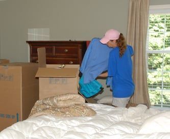 Two people in a bedroom packing boxes. One wears a pink hat and a blue shirt.