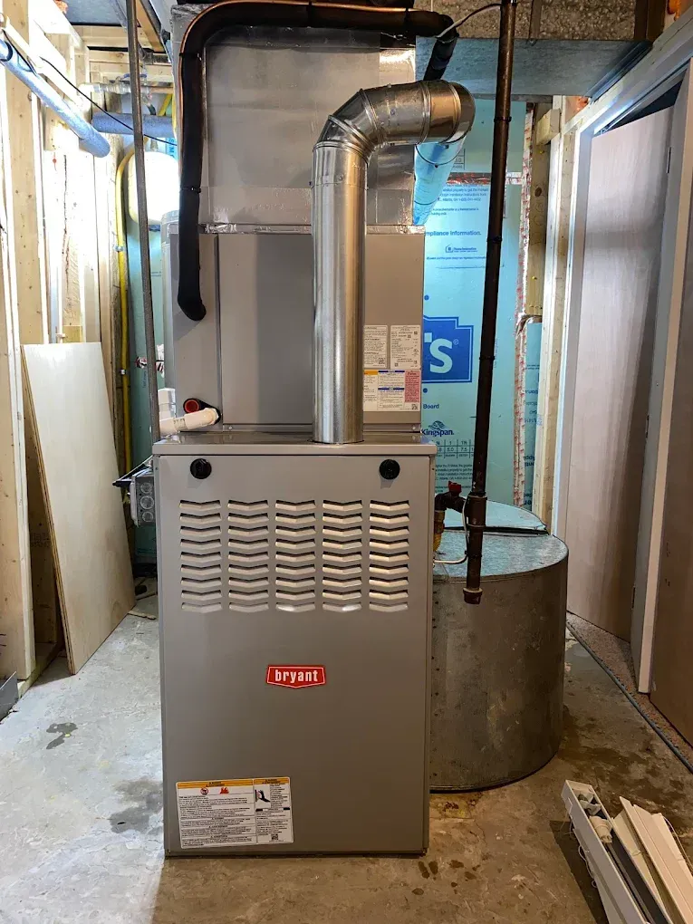 A gray Bryant furnace stands in a utility room next to a metal water heater, with exposed wooden wall studs visible.
