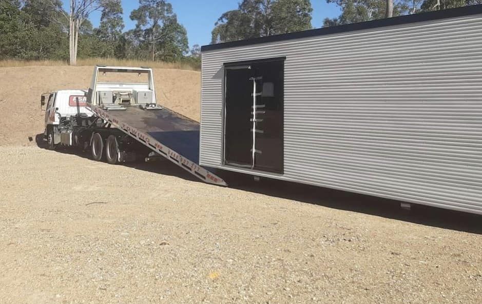 A Large Towing Truck is Loading a Cabin Sized Container on a Dirt Field — ASAP Towing Pty Ltd in Urangan, QLD