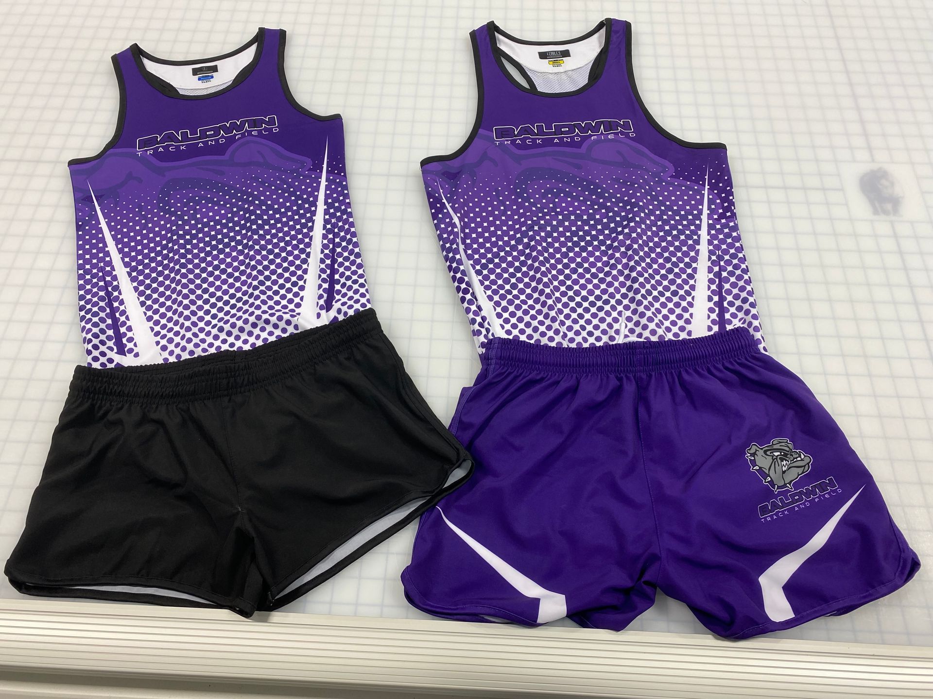 Two track and field uniforms: one black and one purple, with matching purple and white tank tops and shorts.