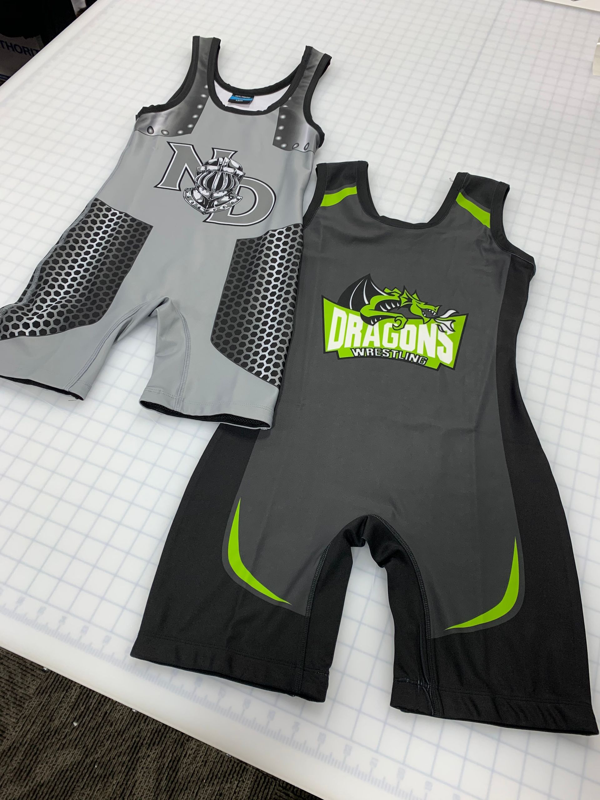 Two grey and black wrestling singlets with logos on a white surface. One has silver accents, the other has green.