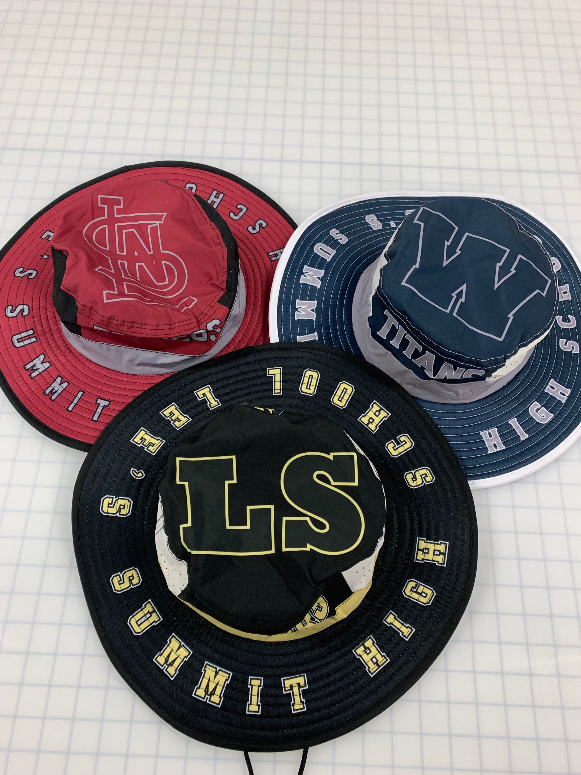 Three sun hats, red, navy, and black, with sports team logos and text around the brim.