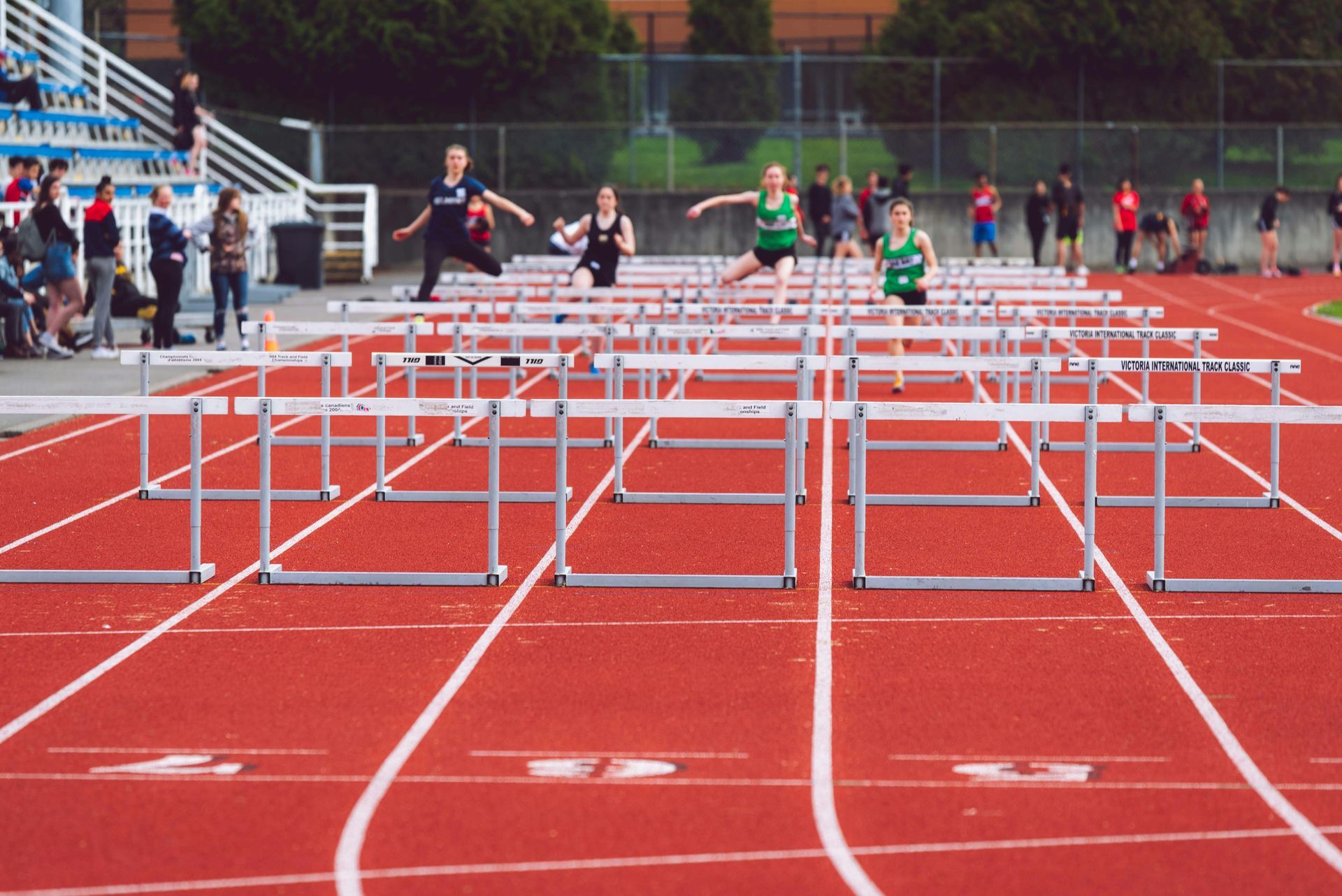 Track athletes in hurdles race on a red track; hurdles lined up.