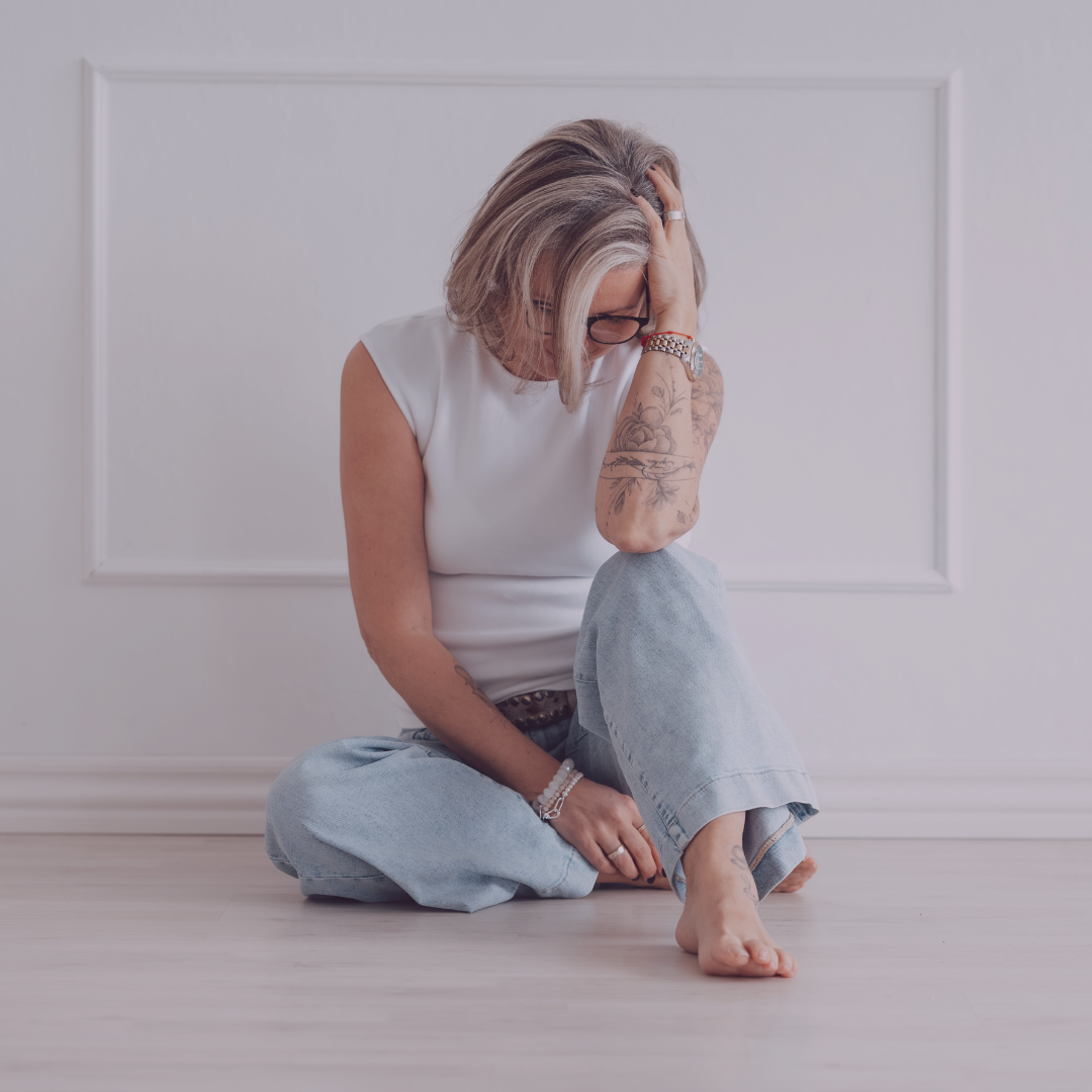 Frau vr weißer Wand, mit blauer Hose und weißem Shirt, schaut nach unten in Trauer.