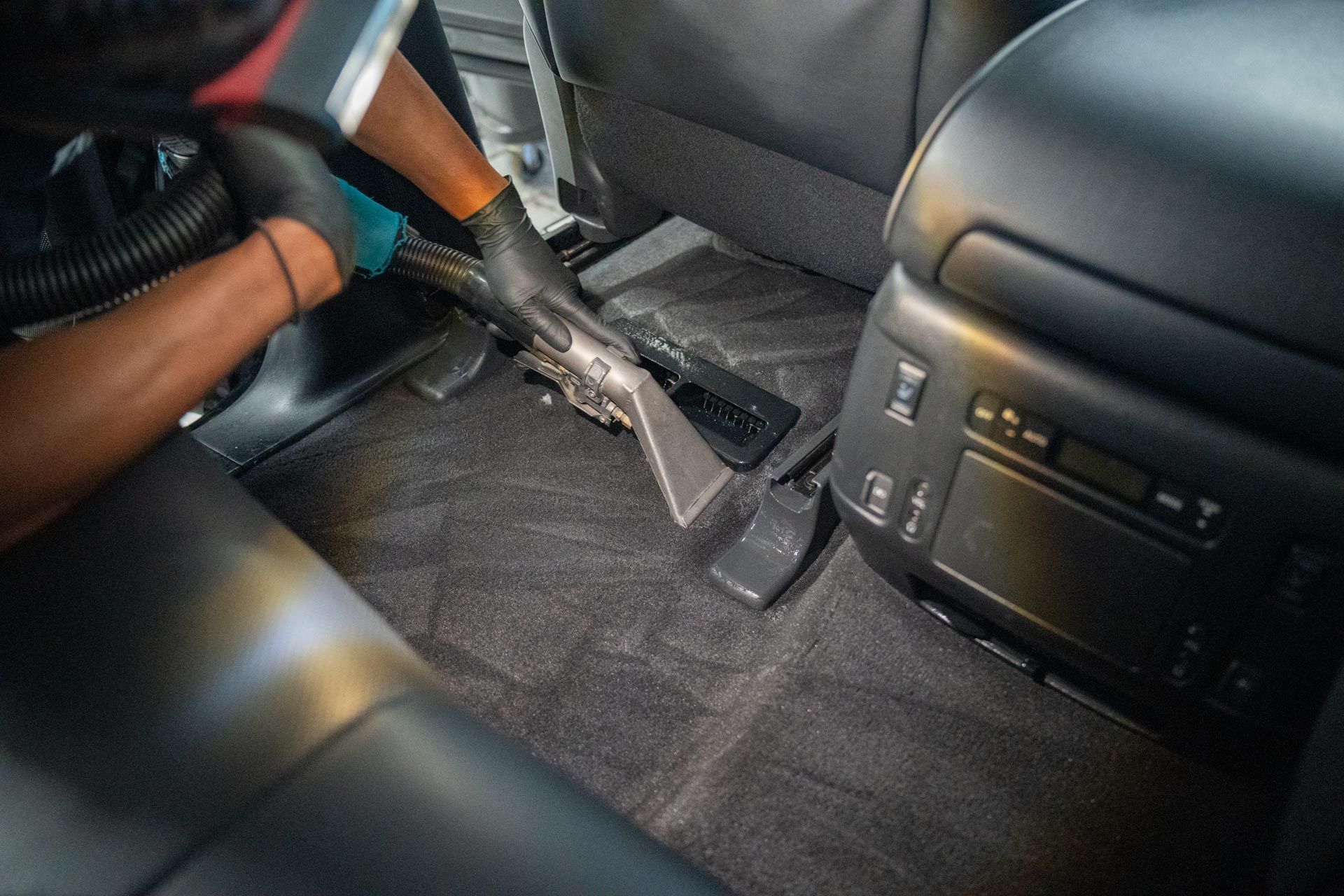 Person vacuuming the interior of a car, focusing on the carpet between seats. Black glove visible.