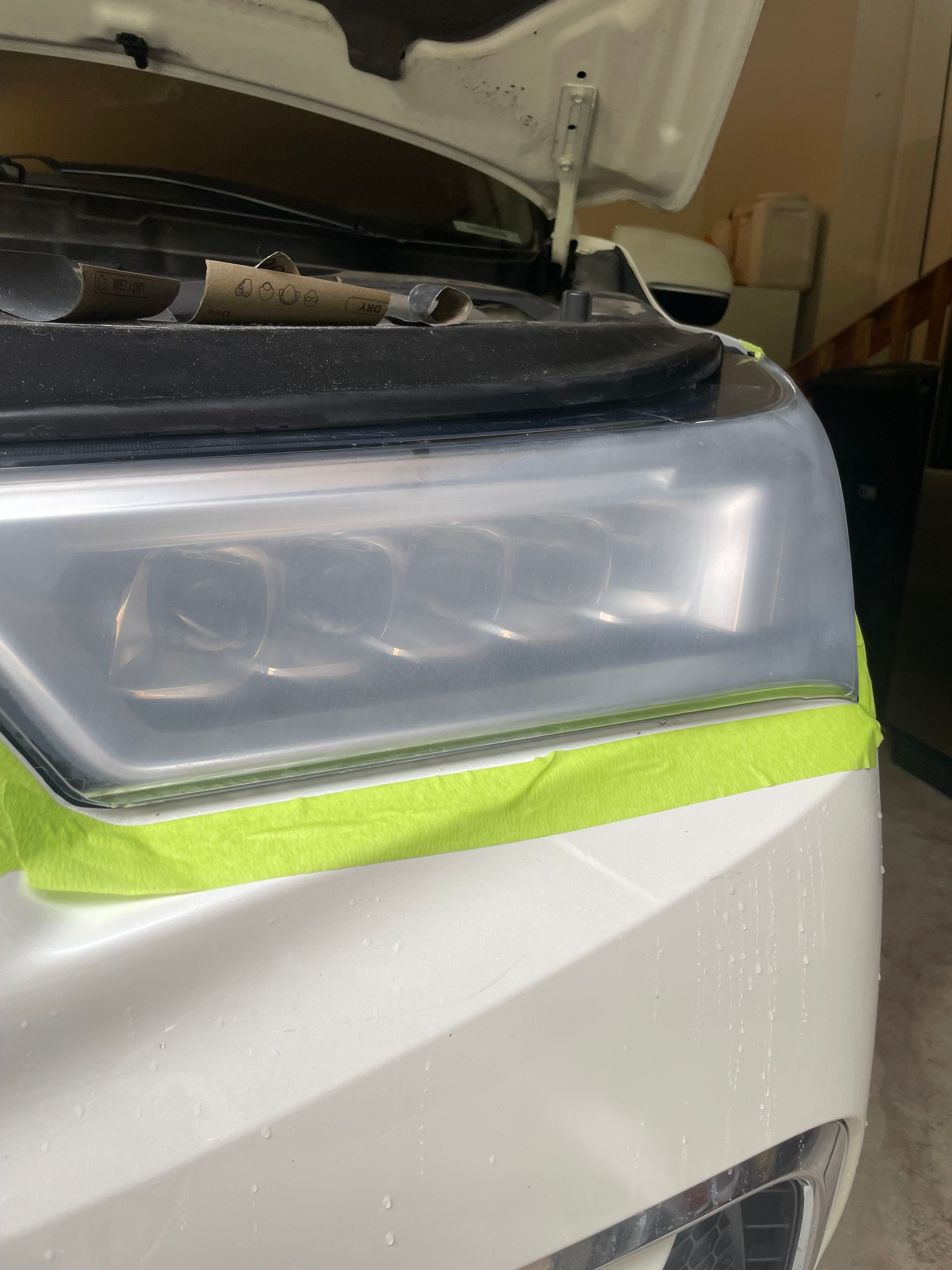 White car headlight being cleaned; taped off with green tape; hood open.