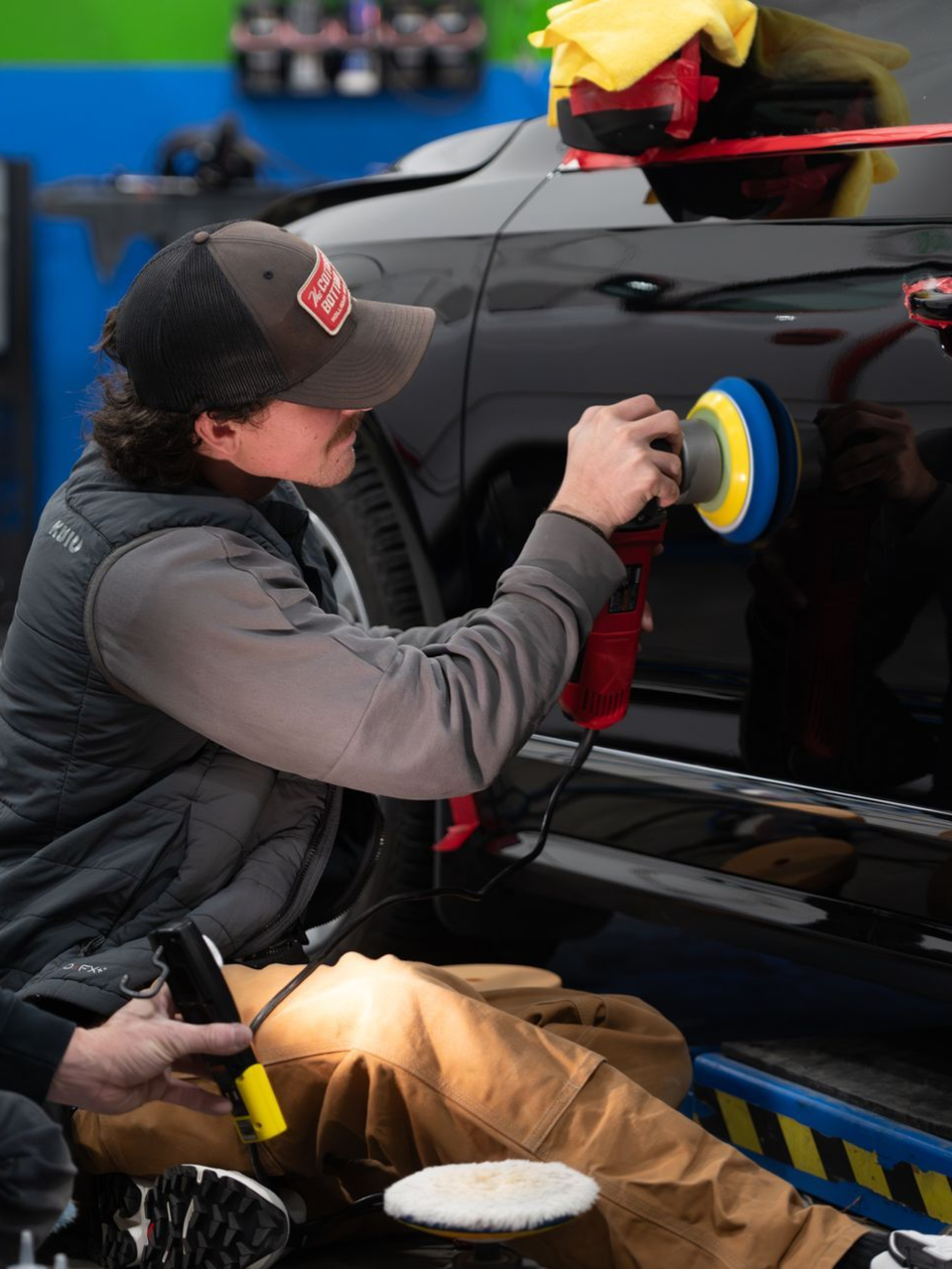 Black car being polished, illuminated by bright lights. A buffer and a bucket are visible.