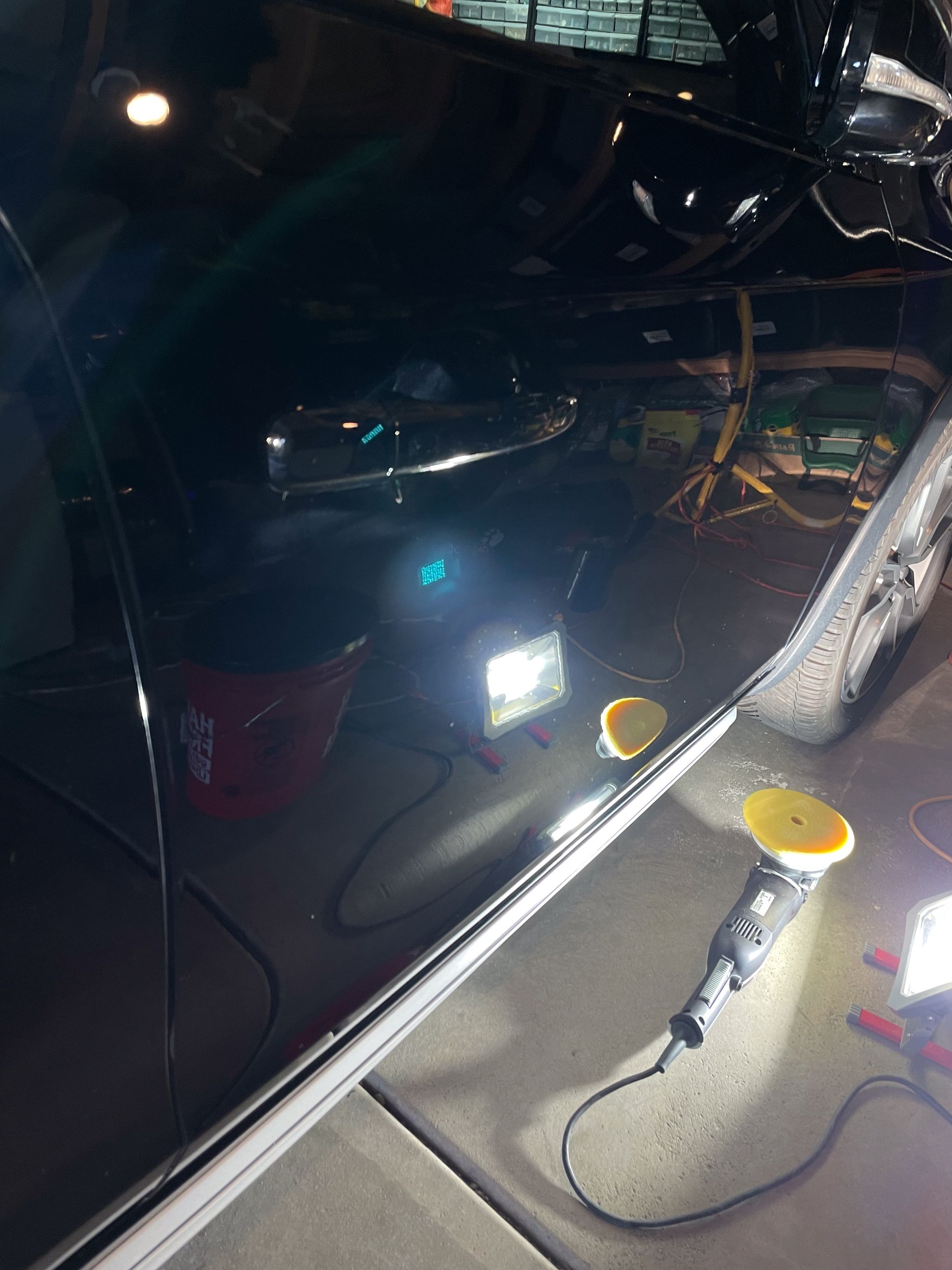 Black car being polished, illuminated by bright lights. A buffer and a bucket are visible.